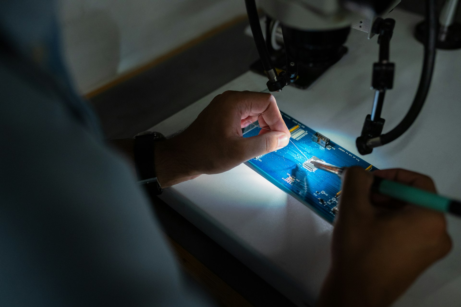 microsoldering person holding a micro soldering iron in one hand and tin in the other soldering a printed circuit board under a microscope